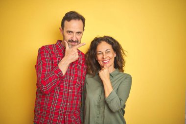 Izole sarı arka plan üzerinde Güzel orta yaş çift çapraz kolları ve çene üzerinde kaldırdı el ile gülümseme ile kameraya emin bakıyor. Olumlu düşünme.