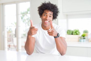 Evde oturan rahat beyaz tişört giyen genç afro-amerikan adam mutlu ve komik yüzü ile kameraya parmaklarını işaret ediyor. İyi enerji ve titreşimler.