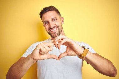 Genç yakışıklı adam sarı izole arka plan üzerinde rahat beyaz t-shirt giyen aşk kalp sembolü ve elleriyle şekil gösteren gülümseyerek. Romantik kavram.