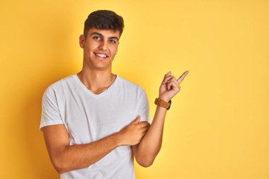 Beyaz tişört giyen genç hintli adam yüzünde büyük bir gülümseme ile izole sarı arka plan üzerinde duran, kameraya bakarak yan el parmağı ile işaret.