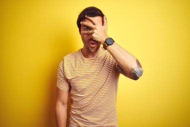 Izole sarı arka plan üzerinde çizgili t-shirt giyen dövme ile genç yakışıklı adam şok el ile yüz ve gözleri kapsayan gözetleme, utanmış ifade ile parmaklarından bakarak.