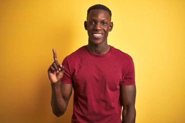 İzole edilmiş sarı arka plan üzerinde duran kırmızı tişört giyen genç afro-amerikan adam, kendinden emin ve mutlu gülümserken bir numaralı parmakla işaret ediyor..