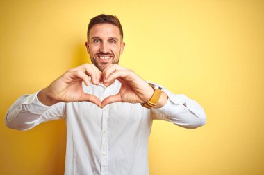 Genç yakışıklı adam sarı izole arka plan üzerinde zarif beyaz gömlek giyen aşk kalp sembolü ve elleriyle şekil gösteren gülümseyerek. Romantik kavram.