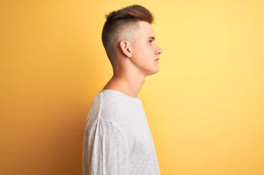 Yan bakarak izole sarı arka plan üzerinde duran beyaz rahat t-shirt giyen genç yakışıklı adam, kendine güvenen gülümseme ile doğal yüz ile profil poz rahat.