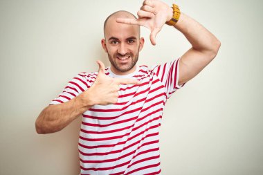 Sakallı genç kel adam beyaz izole arka plan üzerinde rahat çizgili kırmızı t-shirt giyen mutlu yüz ile elleri ve parmakları ile çerçeve yapma gülümseyerek. Yaratıcılık ve fotoğrafçılık konsepti.