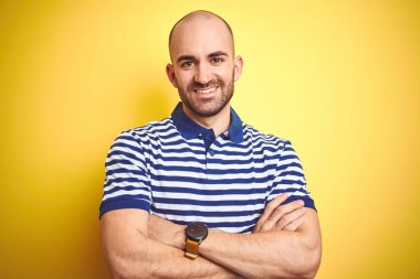Sakallı genç kel adam sarı izole arka plan üzerinde rahat çizgili mavi t-shirt giyen kamera bakarak çapraz kolları ile gülümseyen mutlu yüz. Pozitif kişi.