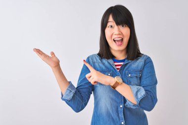 İzole beyaz arka plan üzerinde denim gömlek giyen genç güzel Çinli kadın şaşırttı ve el ile sunarken kameraya gülümseyerek ve parmak ile işaret.