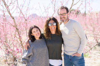 Üç gülümseyen güzel ailesi neşeli ve sarılma şeftali bahçesinde bahar güneşli gün zevk pembe yaprakları ile