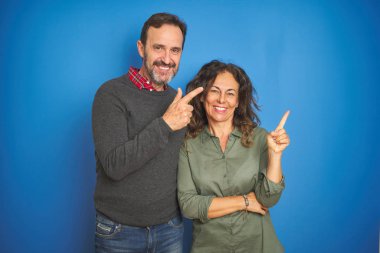 Güzel orta yaş çift birlikte yüzünde büyük bir gülümseme ile izole mavi arka plan üzerinde duran, kameraya bakarak yan el ve parmak ile işaret.