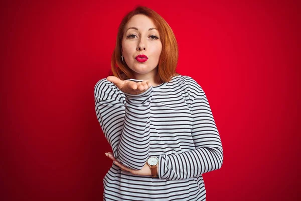 Genç kızıl saçlı kadın kayışlar lacivert gömlek kırmızı izole arka plan üzerinde duran kamera hava güzel ve seksi olmak el iyle bir öpücük üfleme bakarak giyiyor. Aşk ifadesi.