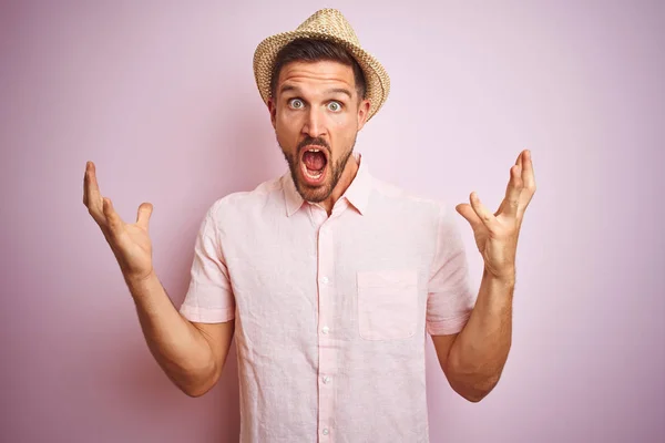 Handsome man wearing hat and summer shirt over pink isolated background celebrating crazy and amazed for success with arms raised and open eyes screaming excited. Winner concept