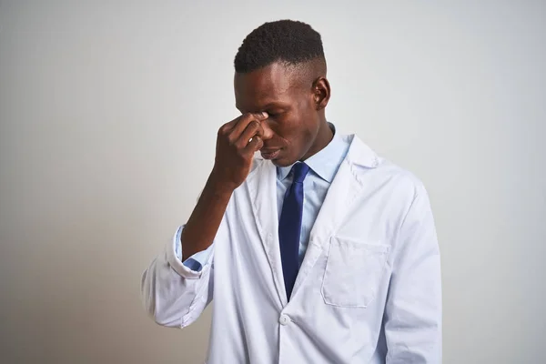Young african american doctor man wearing coat standing over isolated white background tired rubbing nose and eyes feeling fatigue and headache. Stress and frustration concept.