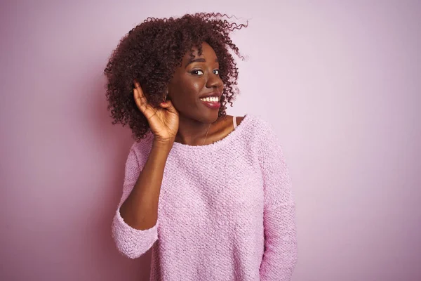 Young african afro woman wearing sweater standing over isolated pink background smiling with hand over ear listening an hearing to rumor or gossip. Deafness concept.