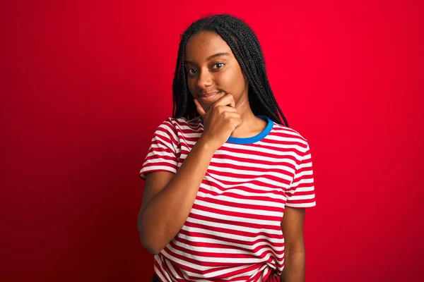 Young african american woman wearing striped t-shirt standing over isolated red background looking confident at the camera smiling with crossed arms and hand raised on chin. Thinking positive.