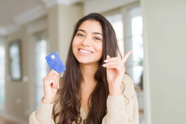 Young woman holding credit card as payment very happy pointing with hand and finger to the side