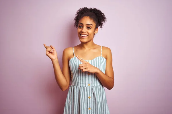 African american woman wearing casual striped dress standing over isolated pink background smiling and looking at the camera pointing with two hands and fingers to the side.