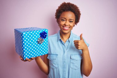 Pembe izole arka plan üzerinde doğum günü hediyesi tutan Afrikalı amerikalı kadın ok işareti yapıyor büyük bir gülümseme ile mutlu, parmakları ile başparmak, mükemmel işareti