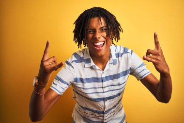Afro amerikan adam dreadlocks izole sarı arka plan üzerinde çizgili gömlek giyen deli ifade elleri ile rock sembolü yapıyor bağırarak. Müzik yıldızı. Ağır konsept.