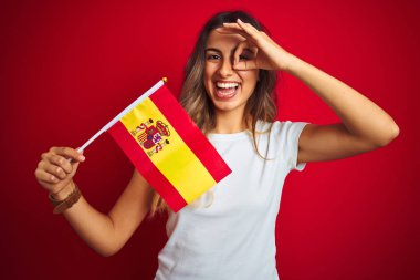 Mutlu yüzü ile kırmızı izole arka plan üzerinde İspanyol bayrağı tutan genç güzel kadın parmakları ile bakarak göz üzerinde el ile ok işareti yapıyor gülümseyerek