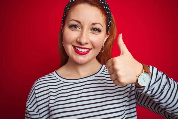 美丽的红发女人穿着条纹海军T恤站在孤立的红色背景做快乐的拇指手势与手 批准表达式 以显示成功的相机 — 图库照片