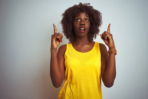 Young african afro woman wearing yellow t-shirt over isolated white background amazed and surprised looking up and pointing with fingers and raised arms.