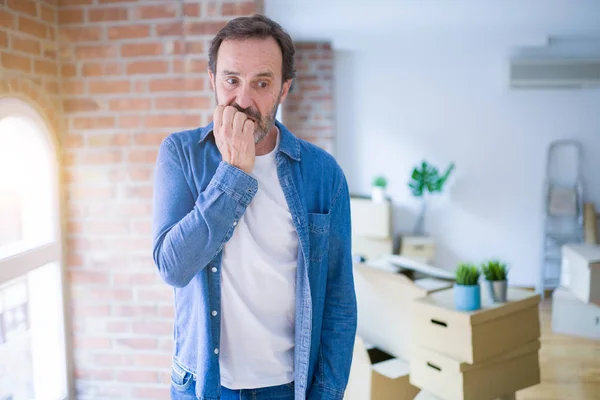 Middle age senior man moving to a new house packing cardboard boxes looking stressed and nervous with hands on mouth biting nails. Anxiety problem.