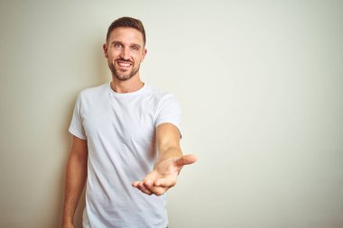 Genç yakışıklı adam izole arka plan üzerinde rahat beyaz t-shirt giyen yardım ve kabul veren neşeli sunan palmiye el gülümseyerek.