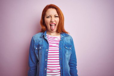 Denim gömlek giyen güzel kızıl saçlı kadın ve izole pembe arka plan üzerinde çizgili t-shirt komik ifade ile mutlu dil yapışmasını. Duygu kavramı.