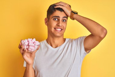 Izole sarı arka plan üzerinde marshmallows ile kase tutan genç Hintli adam baş el ile vurguladı, utanç ve sürpriz yüz ile şok, kızgın ve sinirli. Korku ve hata için üzgün.