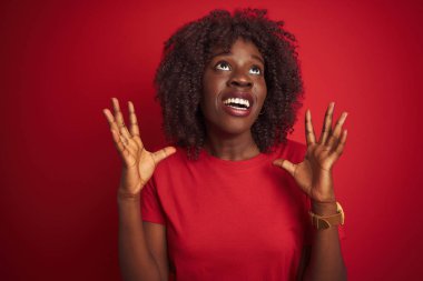 Genç afrikalı afro kadın izole kırmızı arka plan üzerinde duran t-shirt giyen deli ve deli bağırarak ve agresif ifade ve kollar kaldırdı ile bağırıyor. Hayal kırıklığı kavramı.