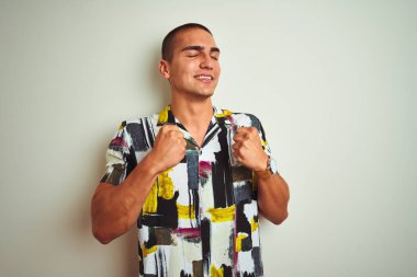 Genç yakışıklı adam beyaz izole arka plan üzerinde yazlık gömlek giyen çok mutlu ve heyecanlı kolları kaldırdı ile kazanan jest yapıyor, gülümseyerek ve başarı için çığlık. Kutlama konsepti.