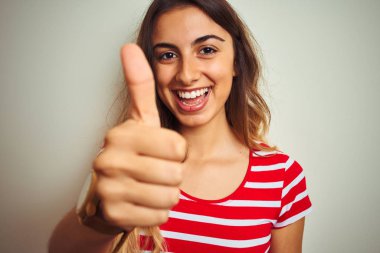 Beyaz izole arka plan üzerinde kırmızı çizgili t-shirt giyen genç güzel kadın ok işareti yapıyor büyük bir gülümseme ile mutlu, parmakları ile başparmak, mükemmel işareti