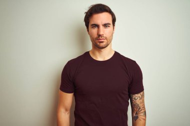 Izole beyaz arka plan üzerinde mor rahat t-shirt giyen dövme ile genç yakışıklı adam yüzünde ciddi ifade ile rahat. Kameraya basit ve doğal bir bakış.