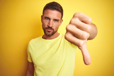 Genç yakışıklı adam sarı izole arka plan üzerinde rahat sarı t-shirt giyen mutsuz ve kızgın gösteren reddi gösteren ve aşağı jest başparmak ile negatif. Kötü ifade.