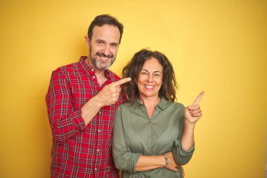 Yüzünde büyük bir gülümseme ile izole sarı arka plan üzerinde Güzel orta yaş çift, kameraya bakarak yan el ve parmak ile işaret.