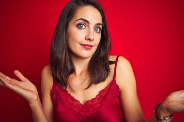 Genç güzel kadın kırmızı izole arka plan üzerinde seksi iç çamaşırı giyen habersiz ve kollar ve eller kaldırdı ile karışık ifade. Şüphe kavramı.