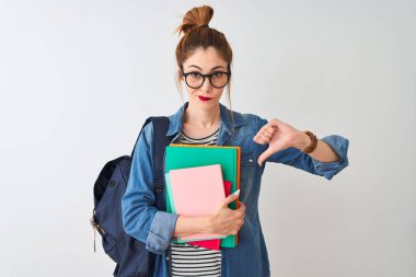 Kızgın yüz ile izole beyaz arka plan üzerinde kitap tutan sırt çantası giyen Redhead öğrenci kadın, başparmak aşağı, ret kavramı ile sevmediğimi gösteren negatif işareti
