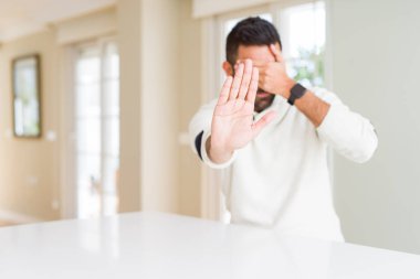 Yakışıklı İspanyol adam evde rahat beyaz kazak giyen elleri ile gözleri kaplayan ve üzgün ve korku ifade ile stop jest yapıyor. Utanmış ve olumsuz kavram.