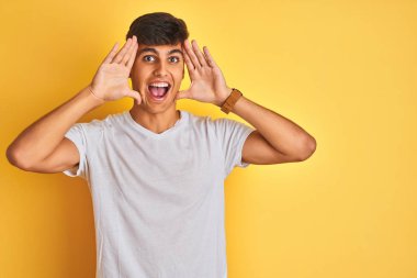 İzole sarı arka plan üzerinde duran beyaz t-shirt giyen genç hintli adam gülümseyen neşeli oynarken elleri yüzünü gösteren bir boo gözetleme. Şaşırttı ve çıktı