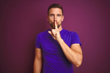 Lila izole arka plan üzerinde rahat mor t-shirt giyen genç adam dudaklarında parmak ile sessiz olmak isteyen. Sessizlik ve gizli kavram.
