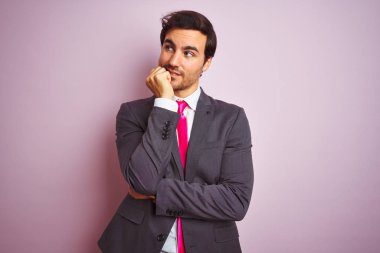 Takım elbise giyen ve kravat takan genç işadamı izole pembe arka plan üzerinde duran stresli ve sinirli ağız tırnakısırma elleri ile bakıyor. Anksiyete sorunu.
