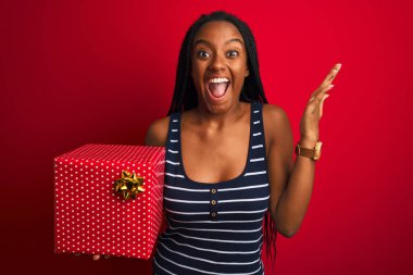 Genç Afrikalı Amerikalı bir kadın elinde doğum günü hediyesi ile kırmızı arka planda mutlu ve heyecanlı duruyor. Kazanan ifade zaferi büyük bir gülümsemeyle kutluyor ve ellerini kaldırıyor.