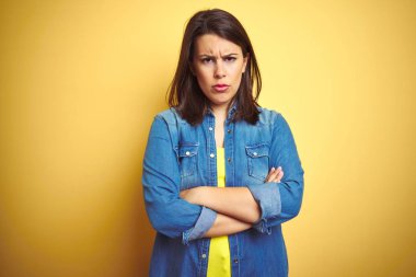 Genç güzel esmer kadın sarı izole arka plan şüpheci ve sinir üzerinde rahat kot ceket giyen, çapraz kolları ile yüzünde onaylamayan ifade. Negatif kişi.