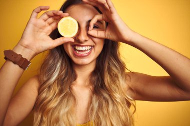 Mutlu yüzü ile sarı izole arka plan üzerinde göz üzerinde bir limon tutan genç güzel kadın parmakları ile bakarak göz üzerinde el ile ok işareti yapıyor gülümseyerek