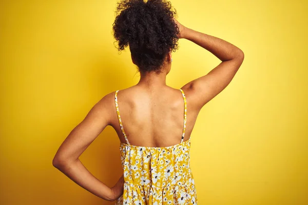 孤立した黄色の背景の上に立つカジュアルな花柄のドレスを着たアフリカ系アメリカ人女性後方に頭に手で疑いを考える — ストック写真