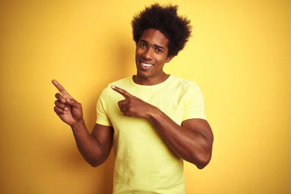 African american man with afro hair wearing t-shirt standing over isolated yellow background smiling and looking at the camera pointing with two hands and fingers to the side.