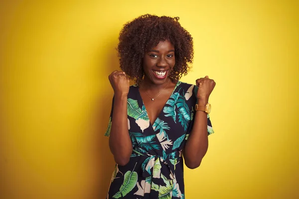 Young african afro woman wearing summer floral dress over isolated yellow background very happy and excited doing winner gesture with arms raised, smiling and screaming for success. Celebration concept.