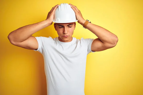 Young handsome man wearing construction helmet over yellow isolated background suffering from headache desperate and stressed because pain and migraine. Hands on head.