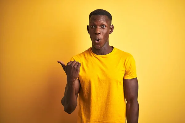 孤立した黄色の背景の上に立つカジュアルなTシャツを着た若いアフリカ系アメリカ人男性が横に手指で指差し 口を開いた表情に驚きました — ストック写真