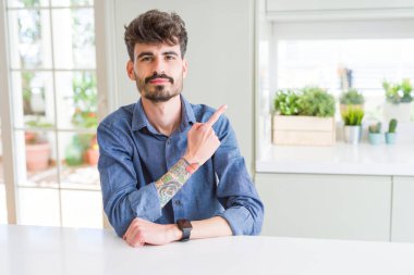 Beyaz masaüzerinde oturan rahat gömlek giyen genç adam yan gösteren el parmağı ile işaret reklam, ciddi ve sakin yüz